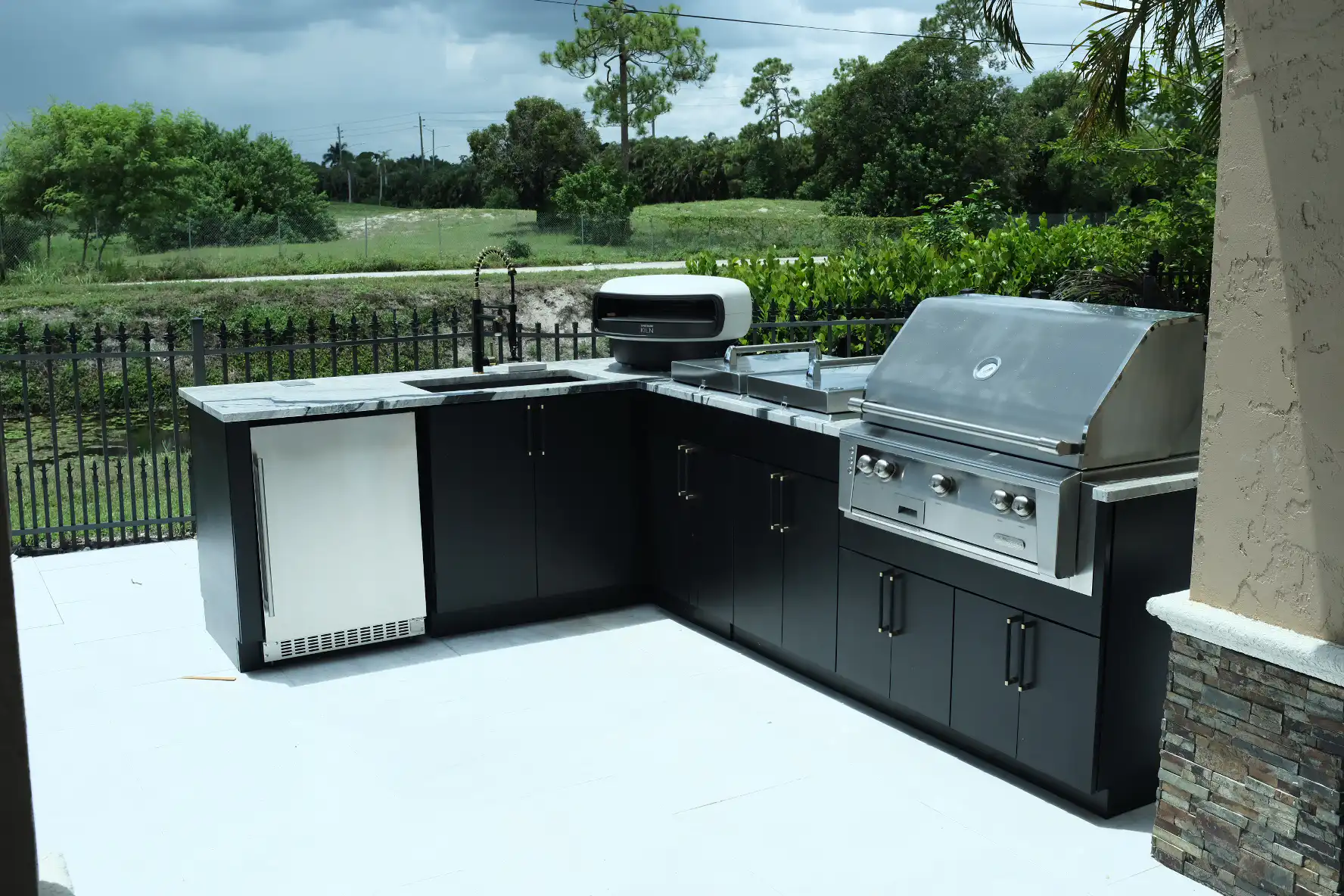 Full L-shaped outdoor kitchen in Lake Worth with Werever black cabinets, Alfresco grill, Everdure pizza oven, Azure fridge, and Eclipse granite countertops