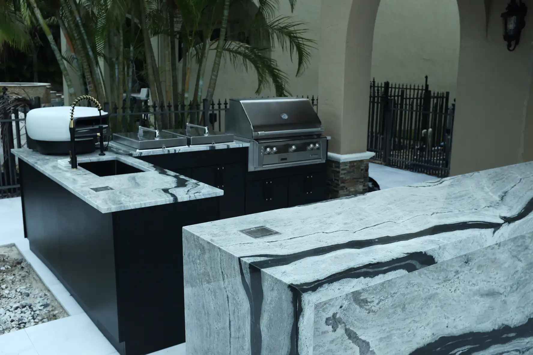 Custom outdoor kitchen with Eclipse granite waterfall bar table, Werever black cabinets, Alfresco grill, and Everdure pizza oven in Lake Worth Florida
