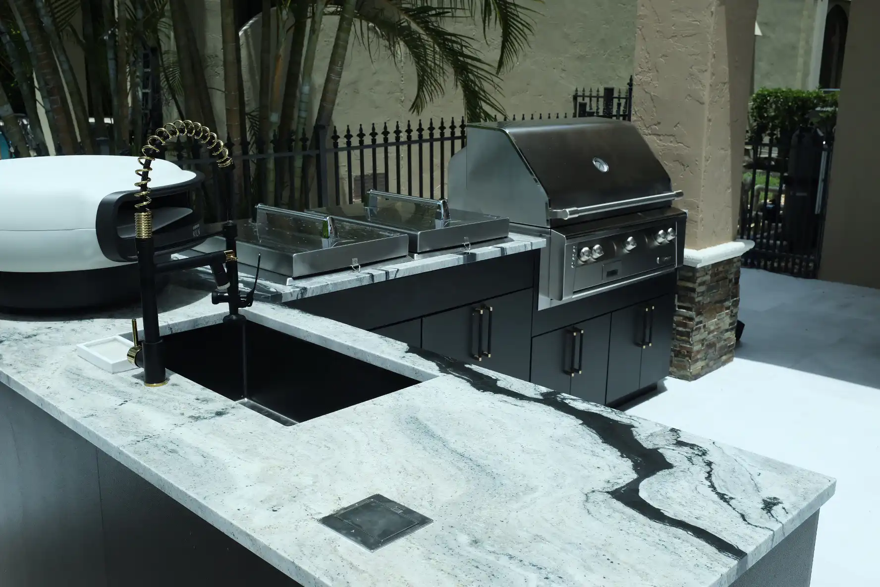 Close-up of Eclipse granite countertop with black sink, gold knurled faucet, Everdure Kiln pizza oven, Lion double burner, and Alfresco grill in custom outdoor kitchen
