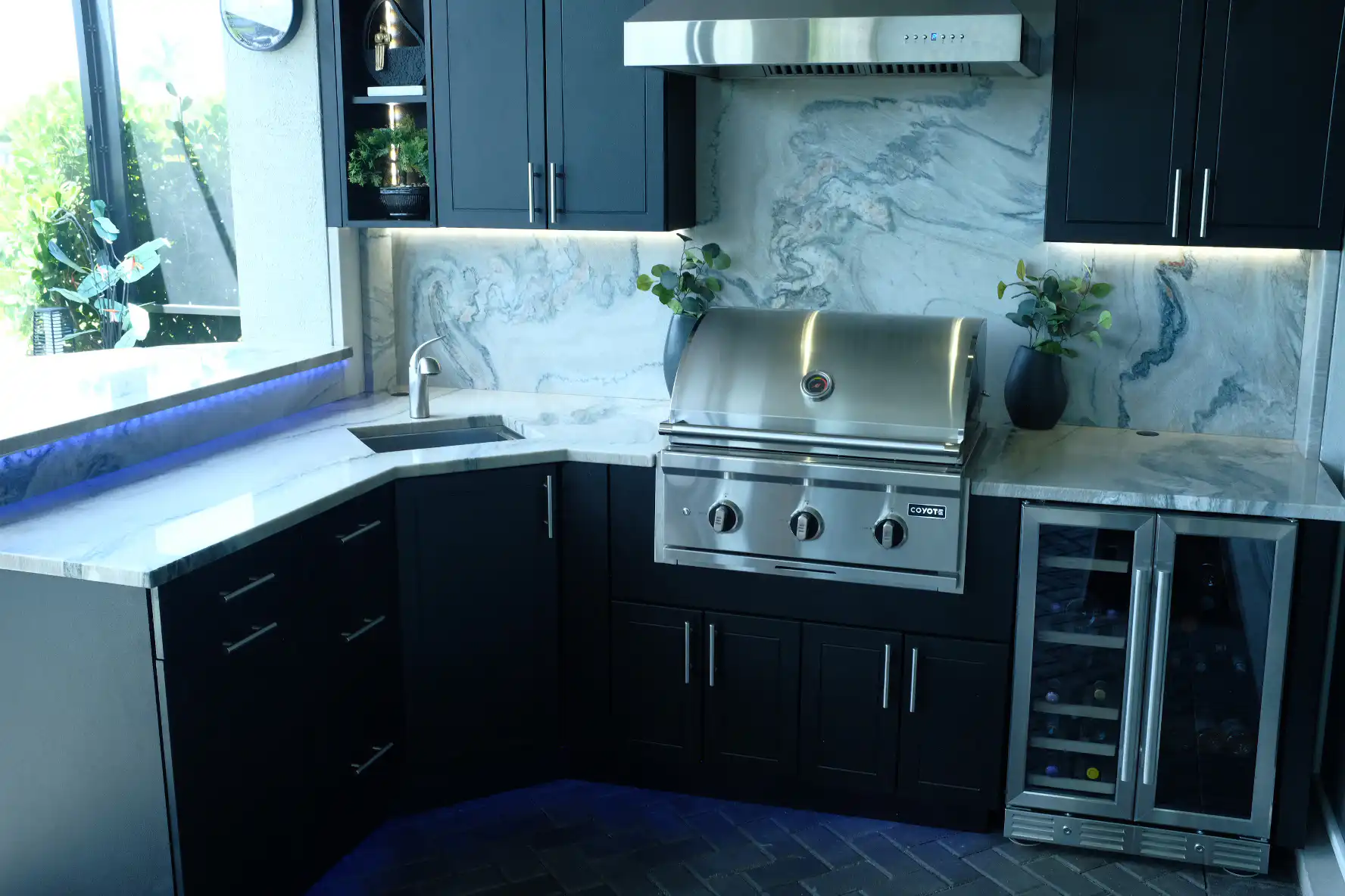 L-shaped outdoor kitchen in Valencia Sound Boynton Beach with Macaubas quartzite, Werever bi-level black cabinets, Coyote grill, and Z-LINE vent hood