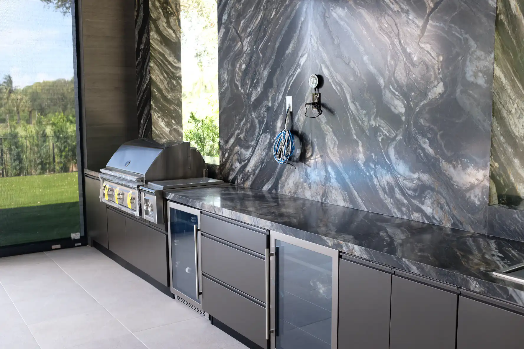 Angled view of Addison Reserve outdoor kitchen highlighting Belvedere quartzite natural stone veining, Lynx infrared grill, Azure refrigerator drawers, and Danver bronze matte cabinetry with retractable screen